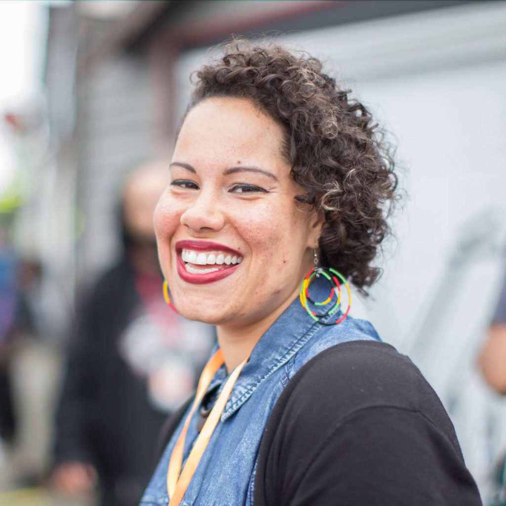 Nikkita Oliver (they-them) smiling in front of a crowd with natural hair and blue jean shirt.