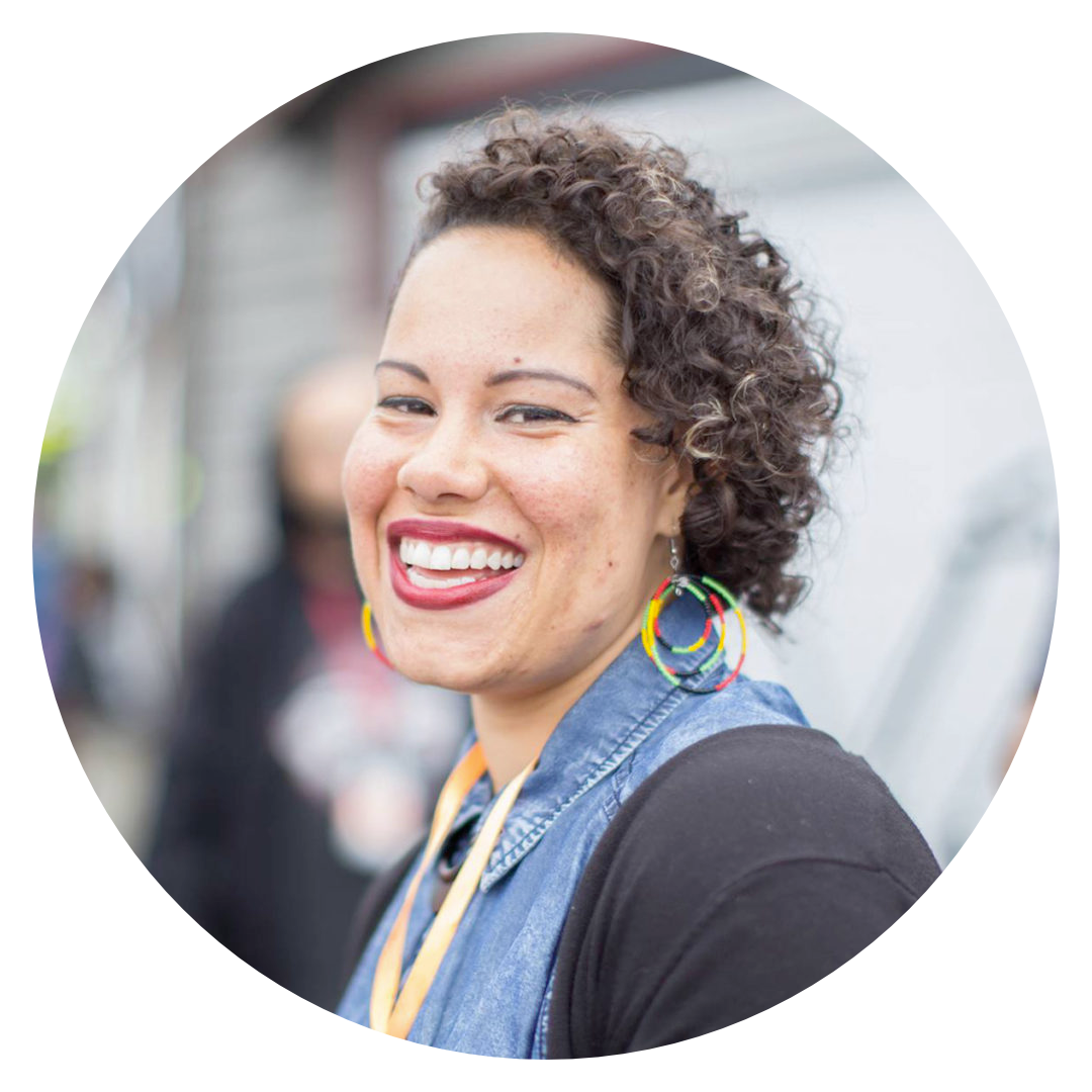 Photo of Nikkita Oliver. They are wearing a blue jean button up shirt, rainbow hoop earrings, a yellow lanyard, and a black cardigan.