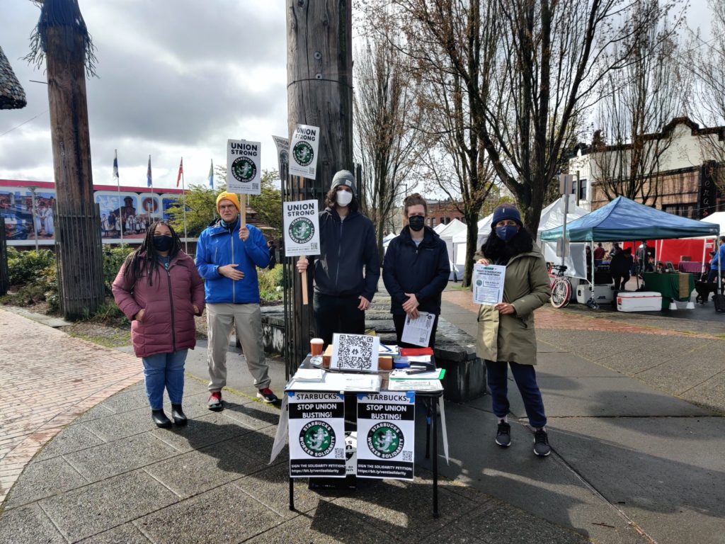 Several Seattle DSA members in front of a table at Ballard farmers market, featuring starbucks solidarity pickets and posters